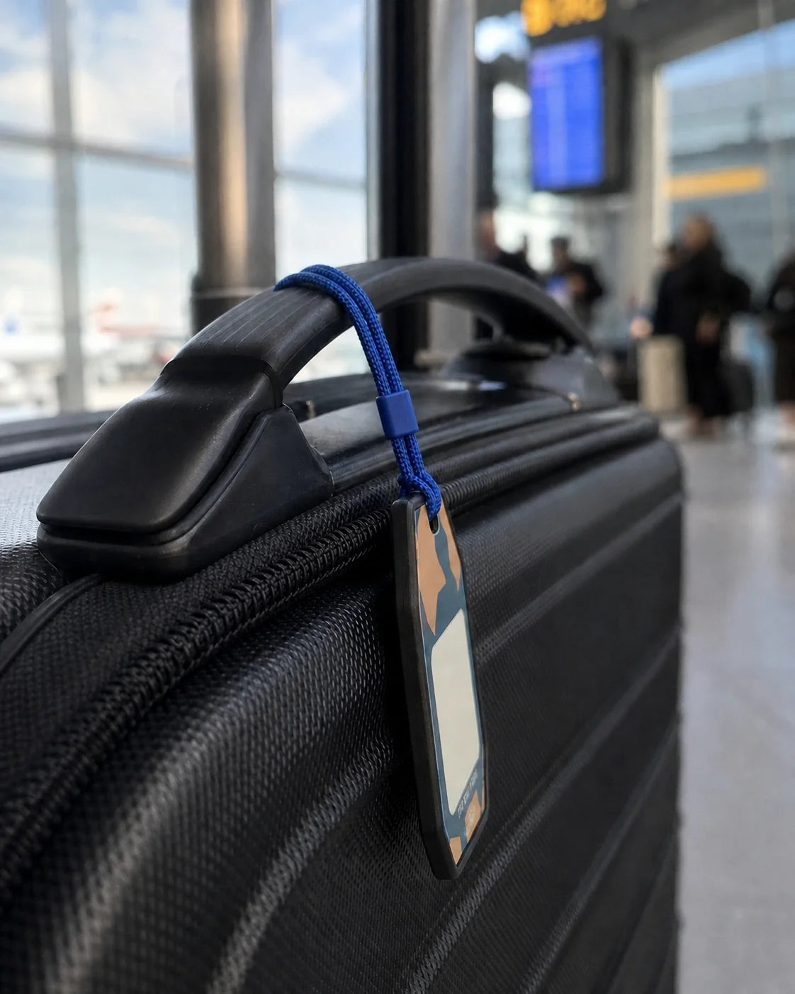 Side-profile airport view of the one-piece 3.2 mm black Tagnook Smart Recovery Tag attached to a carry-on with the supplied blue phone-charm loop.
