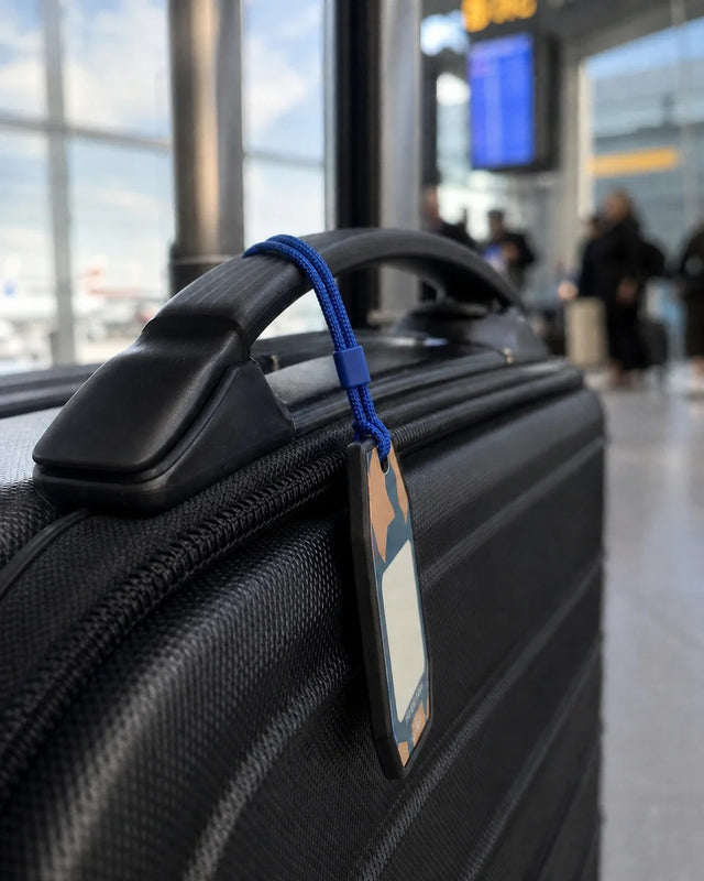 Side-profile airport view of the one-piece 3.2 mm black Tagnook Smart Recovery Tag attached to a carry-on with the supplied blue phone-charm loop.